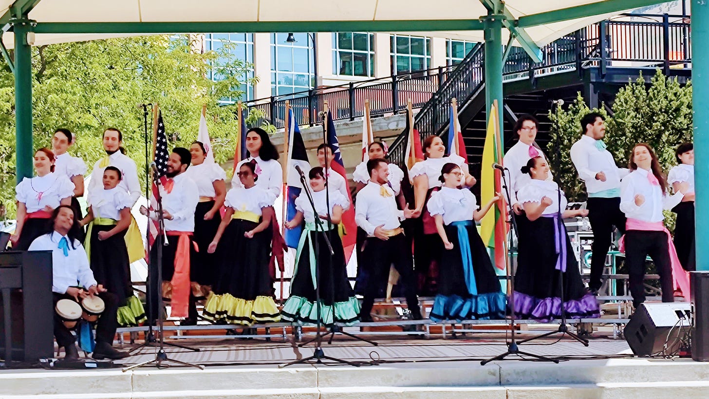 A choir on an outdoor stage, dressed in colorful ethnic-style outfits and dancing along to the music they sing