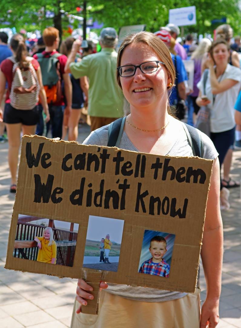 A woman holding a sign at a protest rally. The sign says, "We can't tell them we didn't know." Photos of her with her children are below