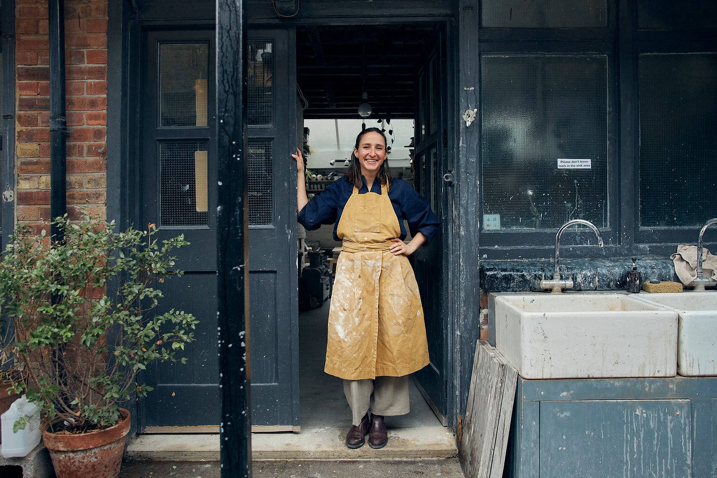 A smiling woman (ceramicist Lucia Ojeco) stands in a doorway in a messy apron. There is an outdoor sink to the right and a plant to the left.