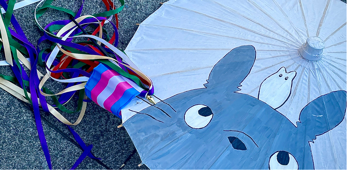cropped photo looking down at a parasol with Totoro painted on it, and a small trans pride flag with a spill of rainbow colored ribbons