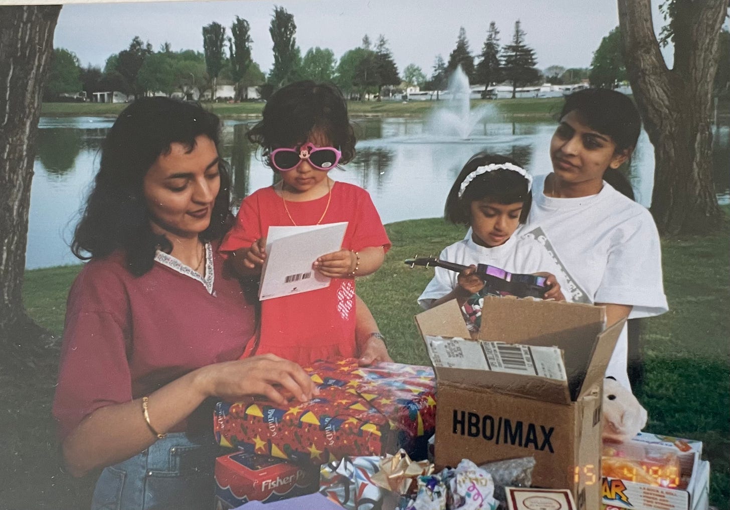 Two mothers with their toddlers. The toddlers play with toys and cards while the mothers look on. They stand in front of a table full of presents and behind them are a pond and trees.