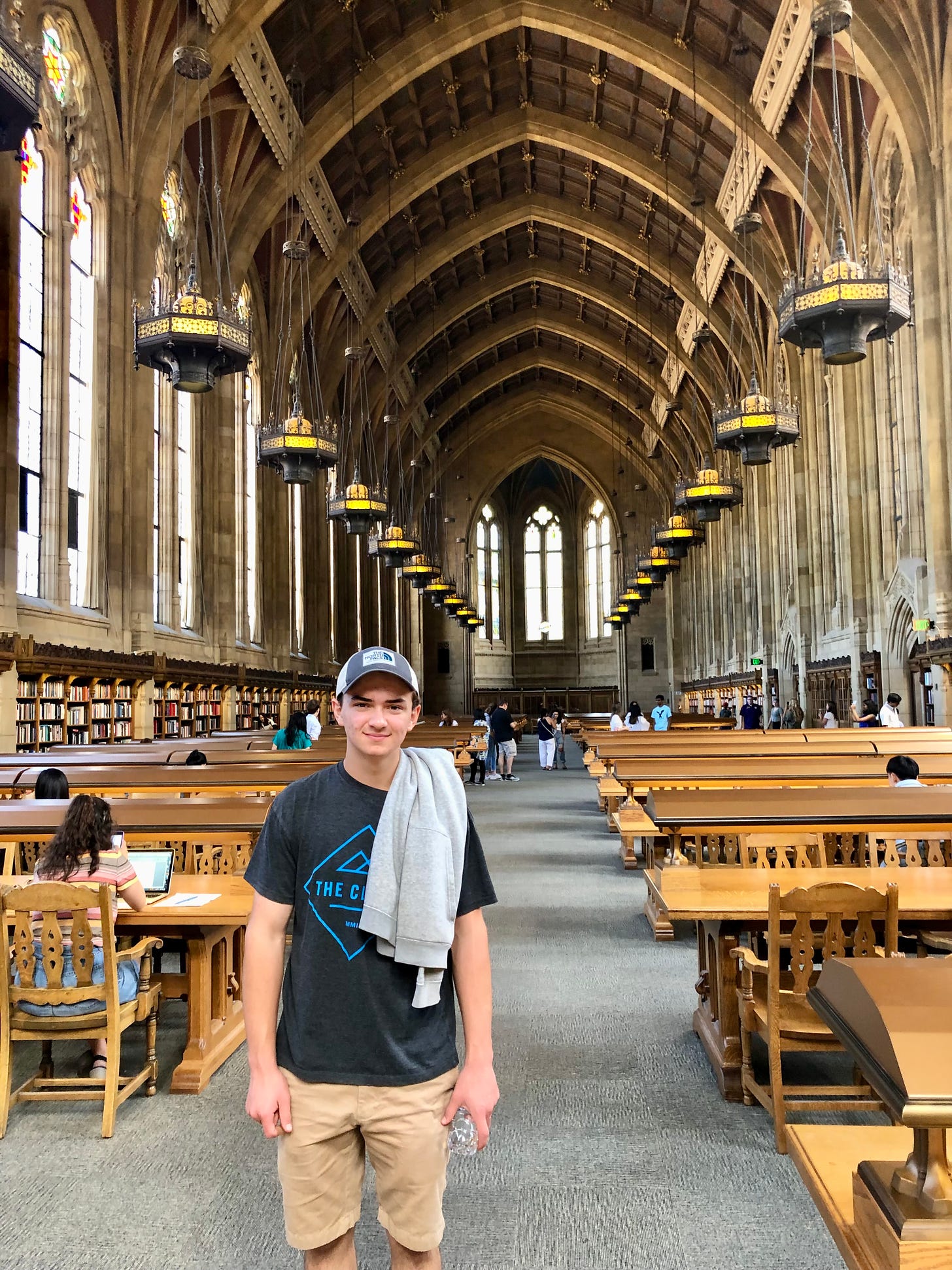 A young white man wearing a t-shirt and basecap stands in a majestic library with gothic vaulted ceilings