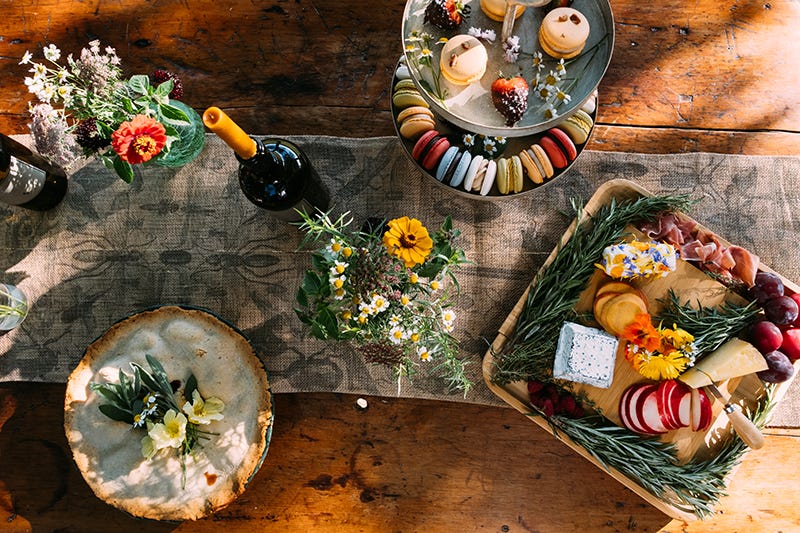A spread of fresh foods and sweets cover a wooden table outdoors with fresh flowers and a bottle of wine
