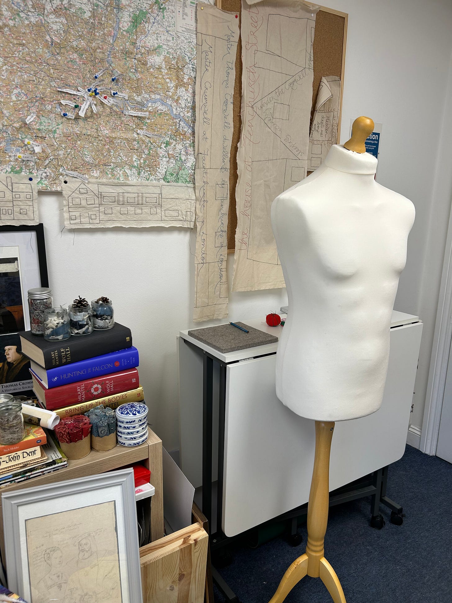 A male dress form in a room that is decorated with a map of London marked up with pins, some strips of embroidered fabric, books about Thomas Cromwell, and portraits 