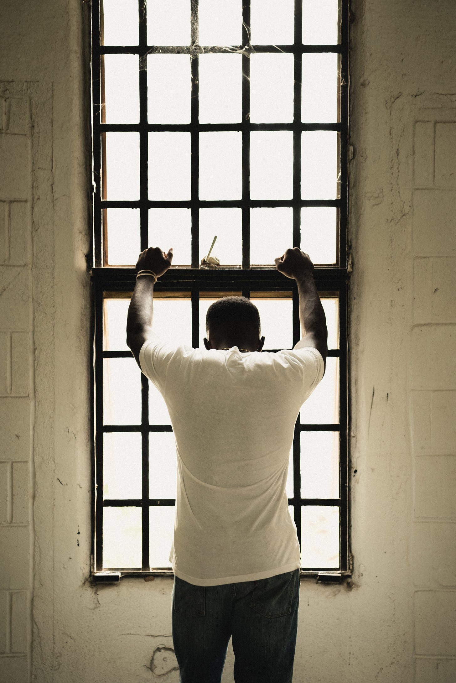 A Black man in a white short-sleeved t-shirt looks out a barred window, his back to the camera.
