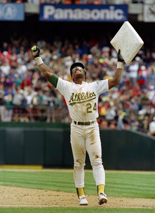 Henderson, wearing an Athletics uniform, stands on a base path with both arms extended and a base in his left hand. He is smiling broadly.