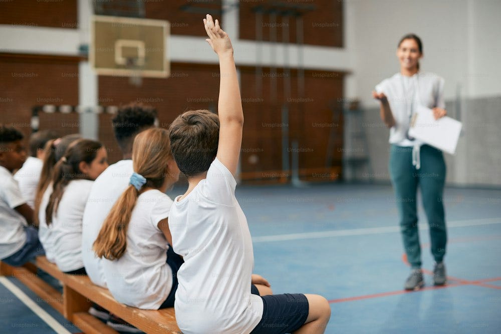 Back view of elementary student raising his arm to answer teacher's question while having PE class at school gym.