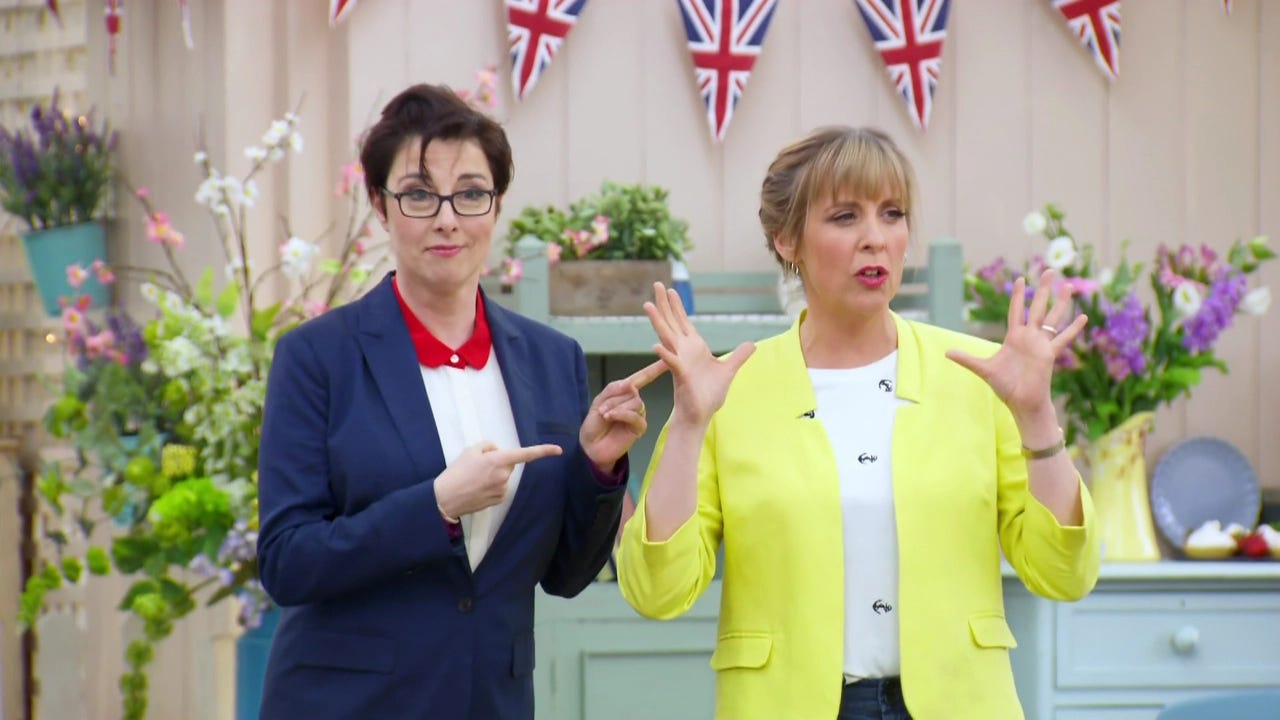 Two white women stand next to each other in a bright, pastel-decorated scene. The woman on the left has short brown hair, wearing a navy blazer over a white shirt. She is pointing with both fingers at the woman on the right, who is a blonde woman smiling with her hands raised and open wide as she talks.