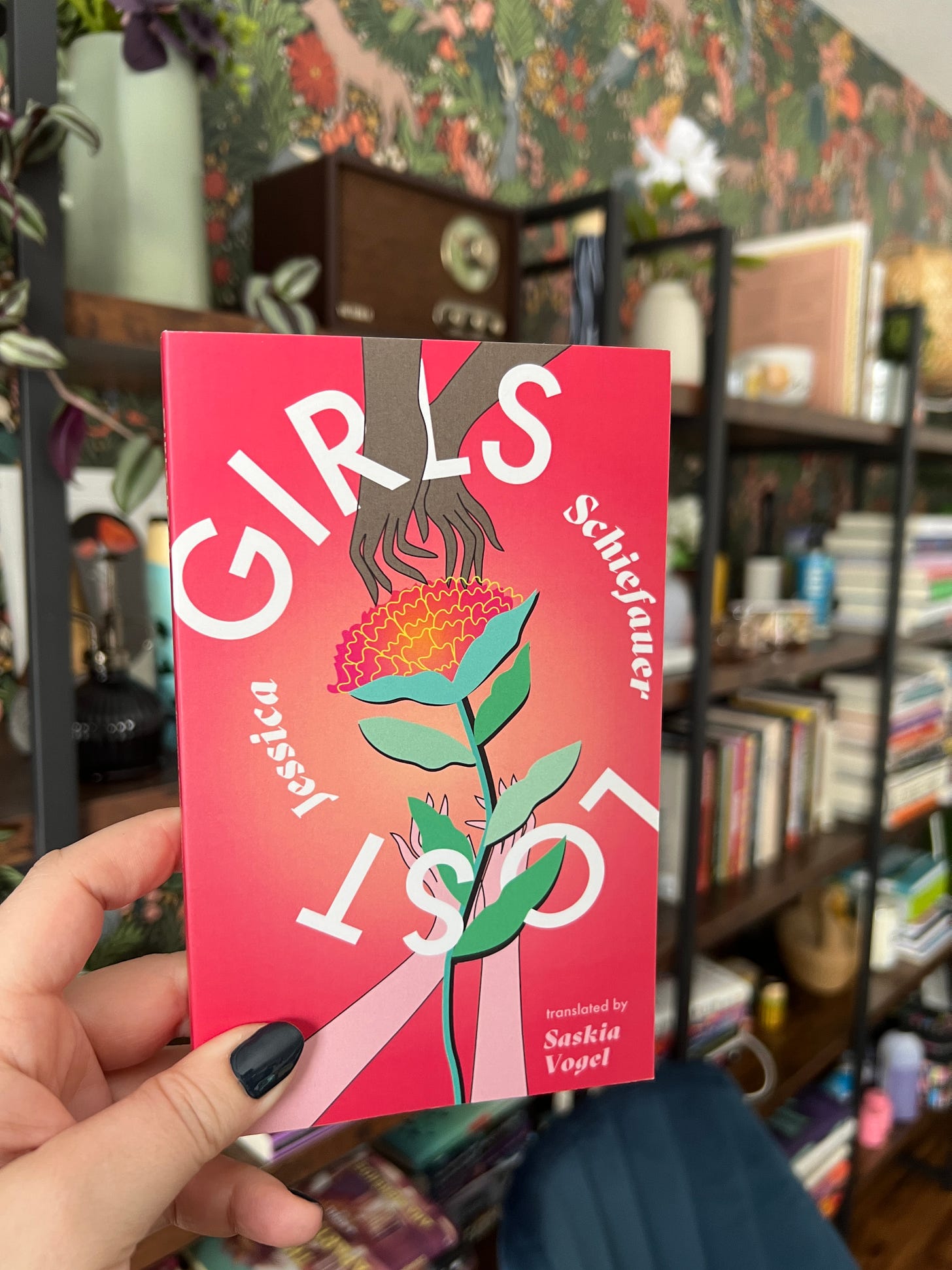 My hand (a white hand) holding a copy of Girls Lost in front of a full bookshelf with plants and books. 