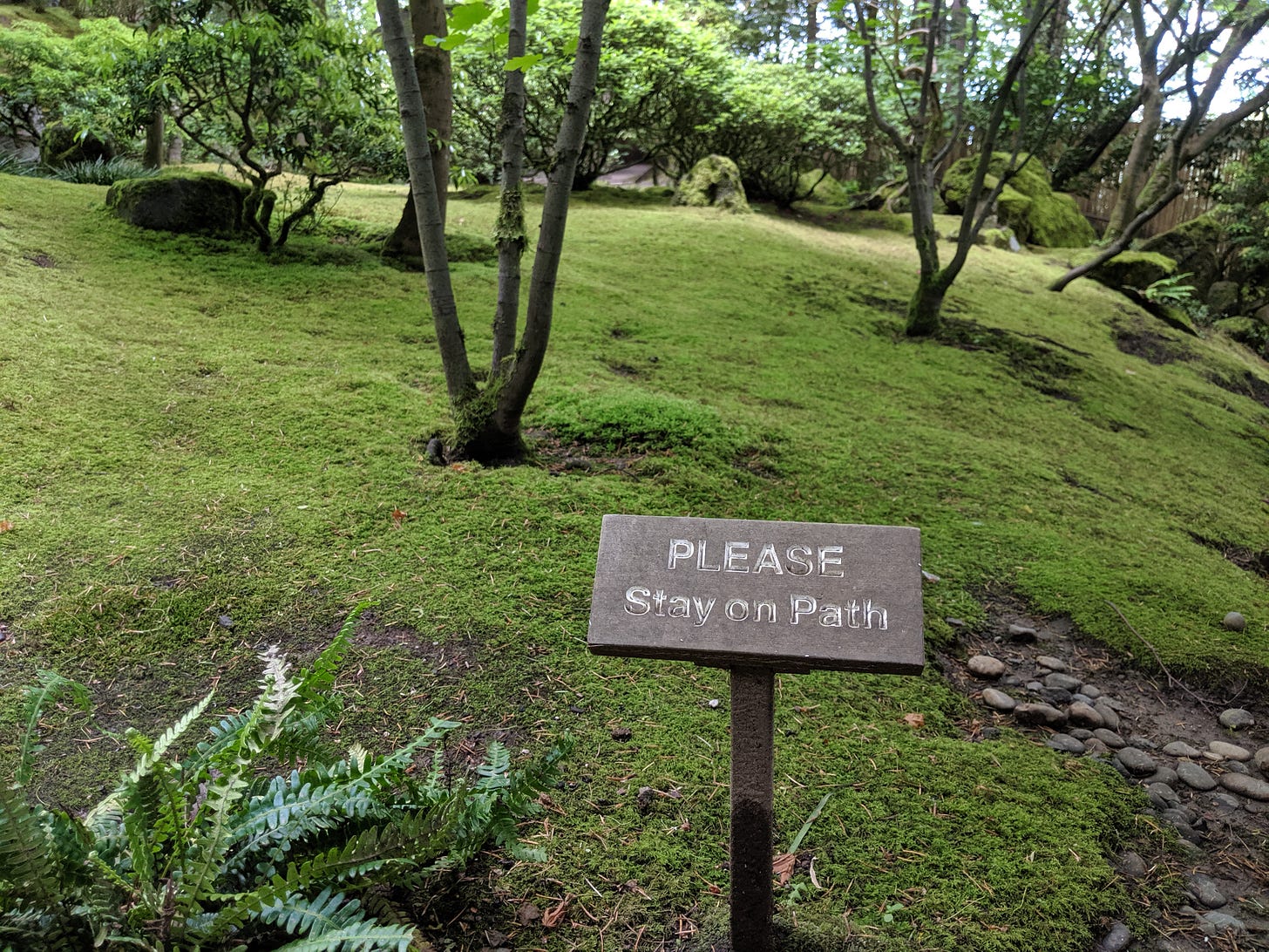 A photo of a sign on a trail that says "Please stay on path", with delicate mosses and small trees in the background