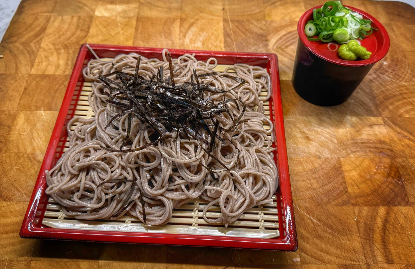 A serving of zarusoba topped with shredded seaweed on a traditional zaru mat. A cup of dipping sauce is beside it, topped with a lid holding green onions and wasabi