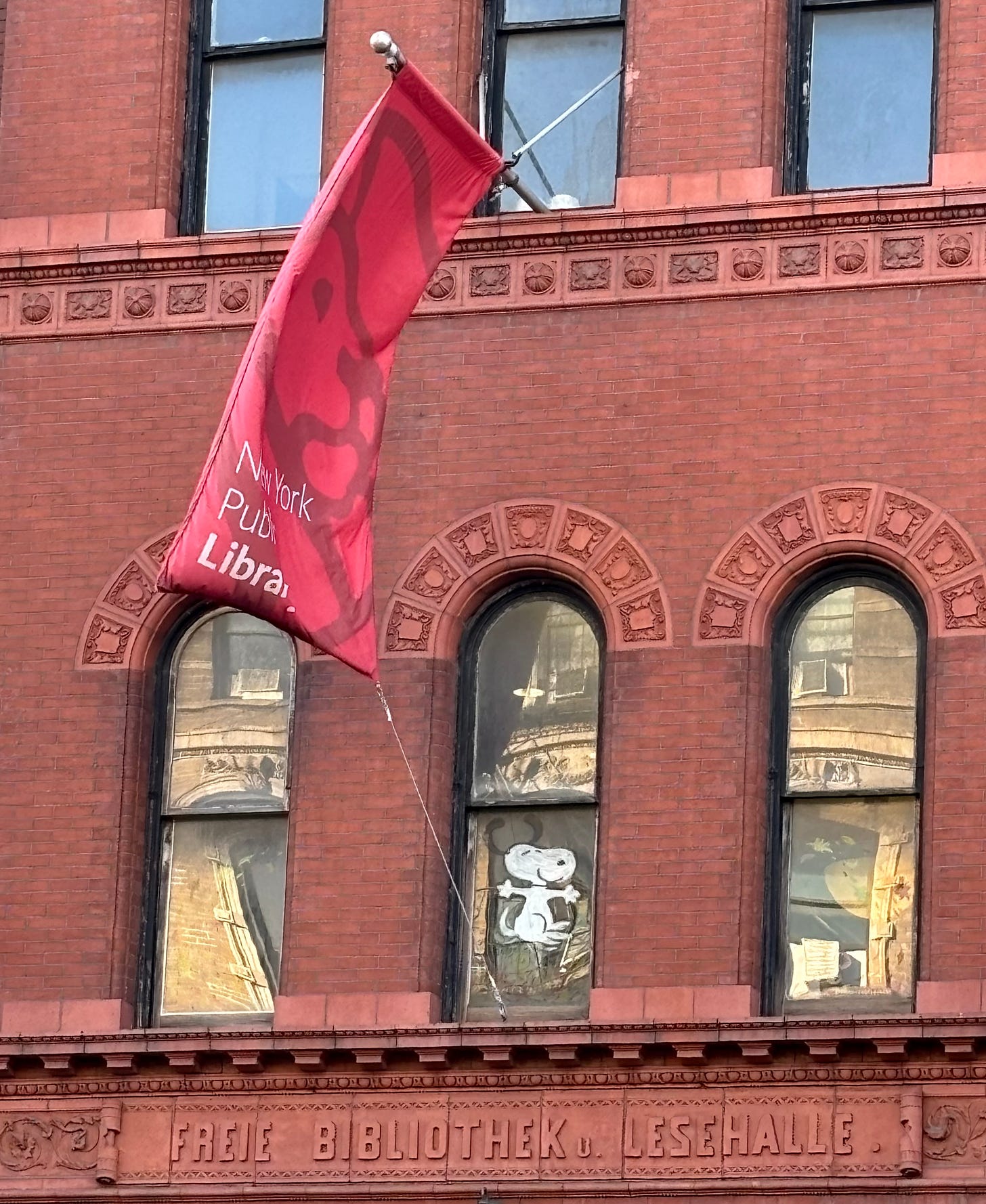 The Ottendorfer NYPL brach. Over the door is engraved FREIE BIBLIOTHEK u. LESEHALLE. A red NYPL flag is blowing in the wind. In the window, someone has painting snoopy. He's really happy you're reading.