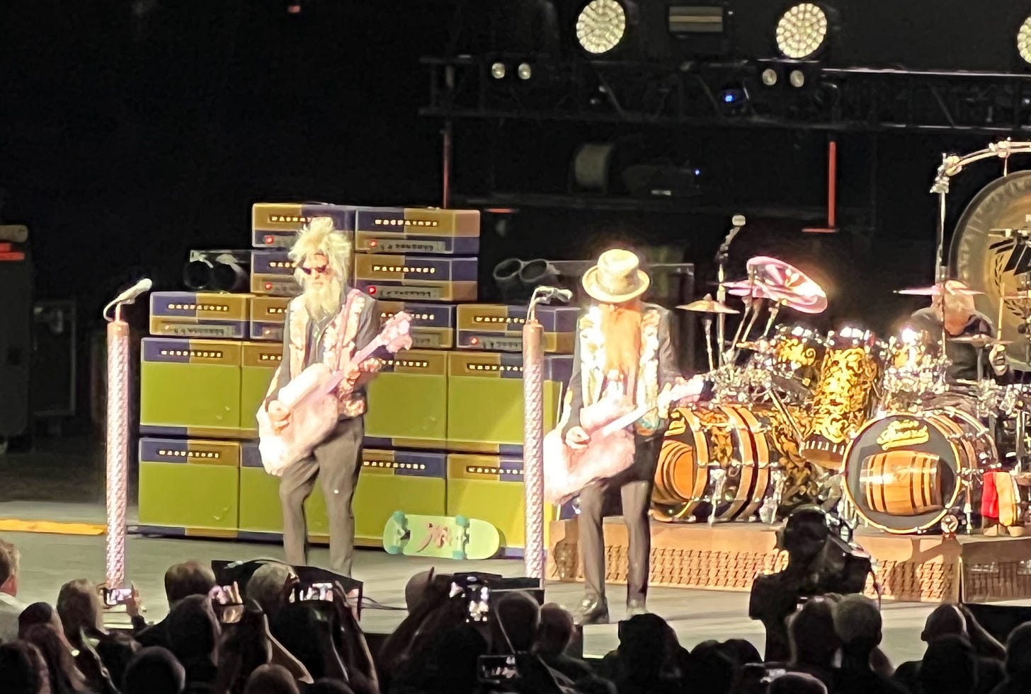 A cell phone photo of ZZ Top playing on stage, with bassist Elwood Francis and guitarist Billy Gibbons in front. Stacks of lime-green amps and a drum kit and drummer Frank Beard are behind. A skateboard is propped against the amps.