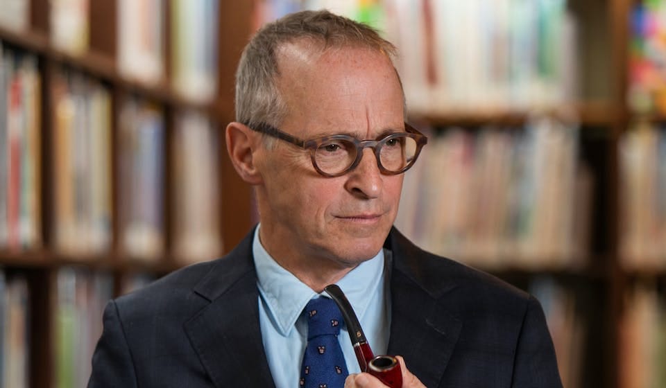 A photograph of David Sedaris, a white man with short grey hair. He is wearing glasses, a button down shirt , blazer and tie and holds a pipe.