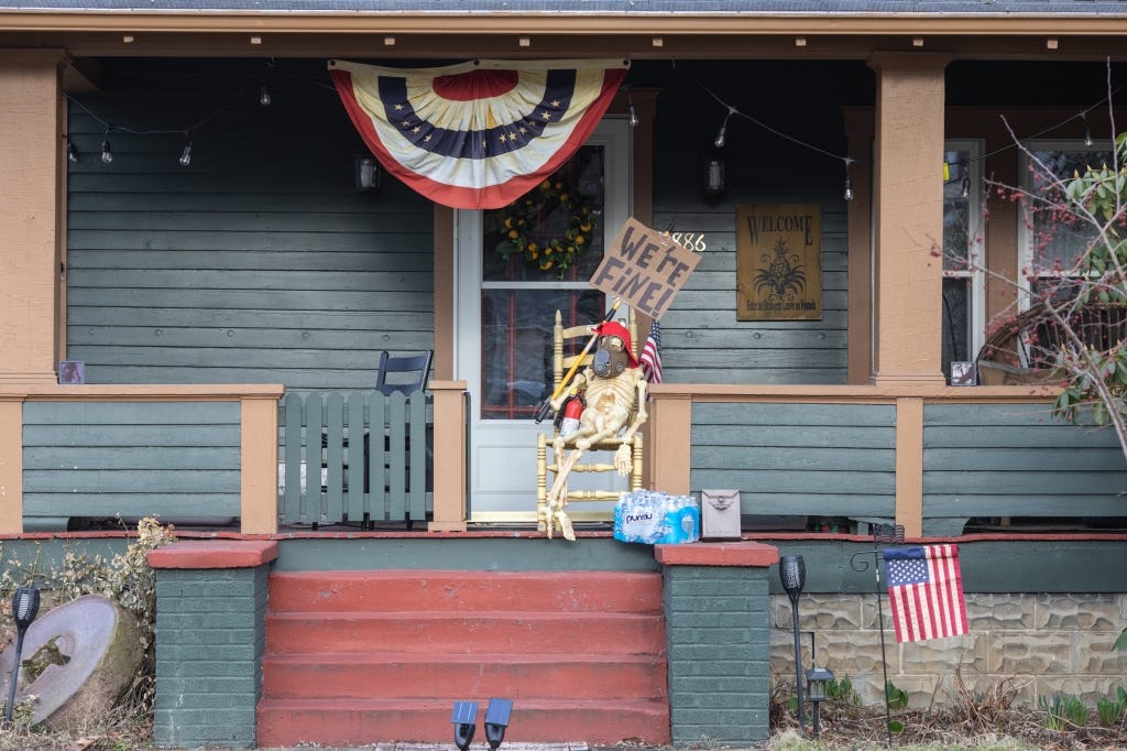 A skeleton with a gas mask and a case of bottled water and a sign reading "We're fine!" sits on the porch of a house in downtown East Palestine.