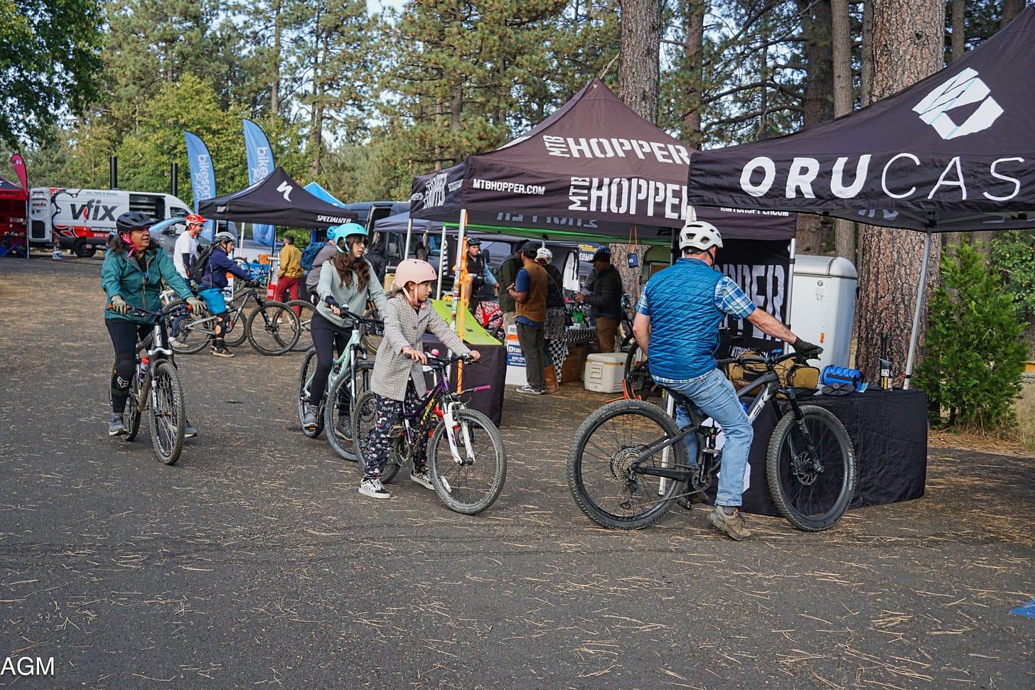 A family of four stop in front of a pop-up tent on their mountain bikes. A mom, elementary and teen daughters, and father in the lead check out bike bags. The tent is the first in a line of vendors set up in front of pine trees.