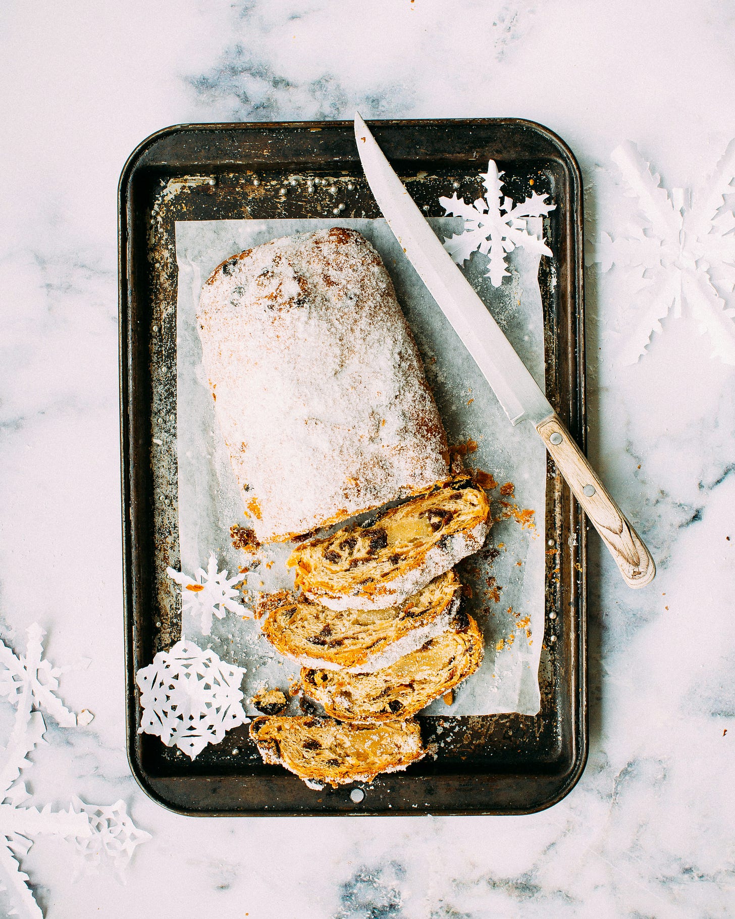 A stollen loaf sits on a well-used baking tray and a sheet of baking parchment, surrounded by paper cut-out snowflakes. The stollen has been cut, and four slices are laid on their sides to show the dried fruit and marzipan inside. A vintage knife sits beside the stollen.