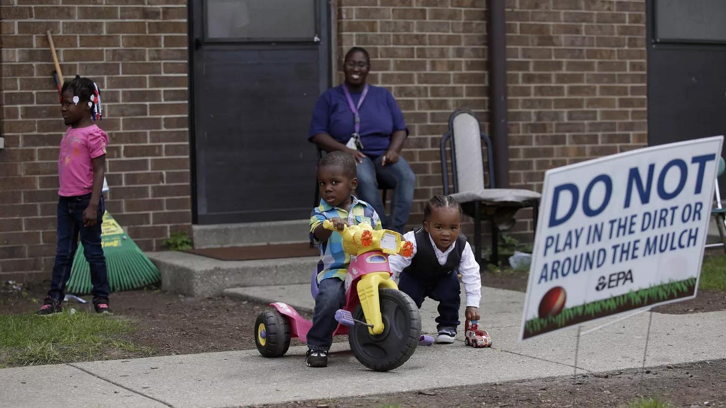 A mother sits outside of her apartment door as her two young sons play, one on a tricycle and the other with a toy car. The mother’s young daughter walks away past a rake leaning against the building. A sign in the dirt reads, “Do not play in the dirt or around the mulch. —EPA.” A mother sits outside of her apartment door as her two young sons play, one on a tricycle and the other with a toy car. The mother’s young daughter walks away past a rake leaning against the building. A sign in the dirt reads, “Do not play in the dirt or around the mulch. —EPA.”