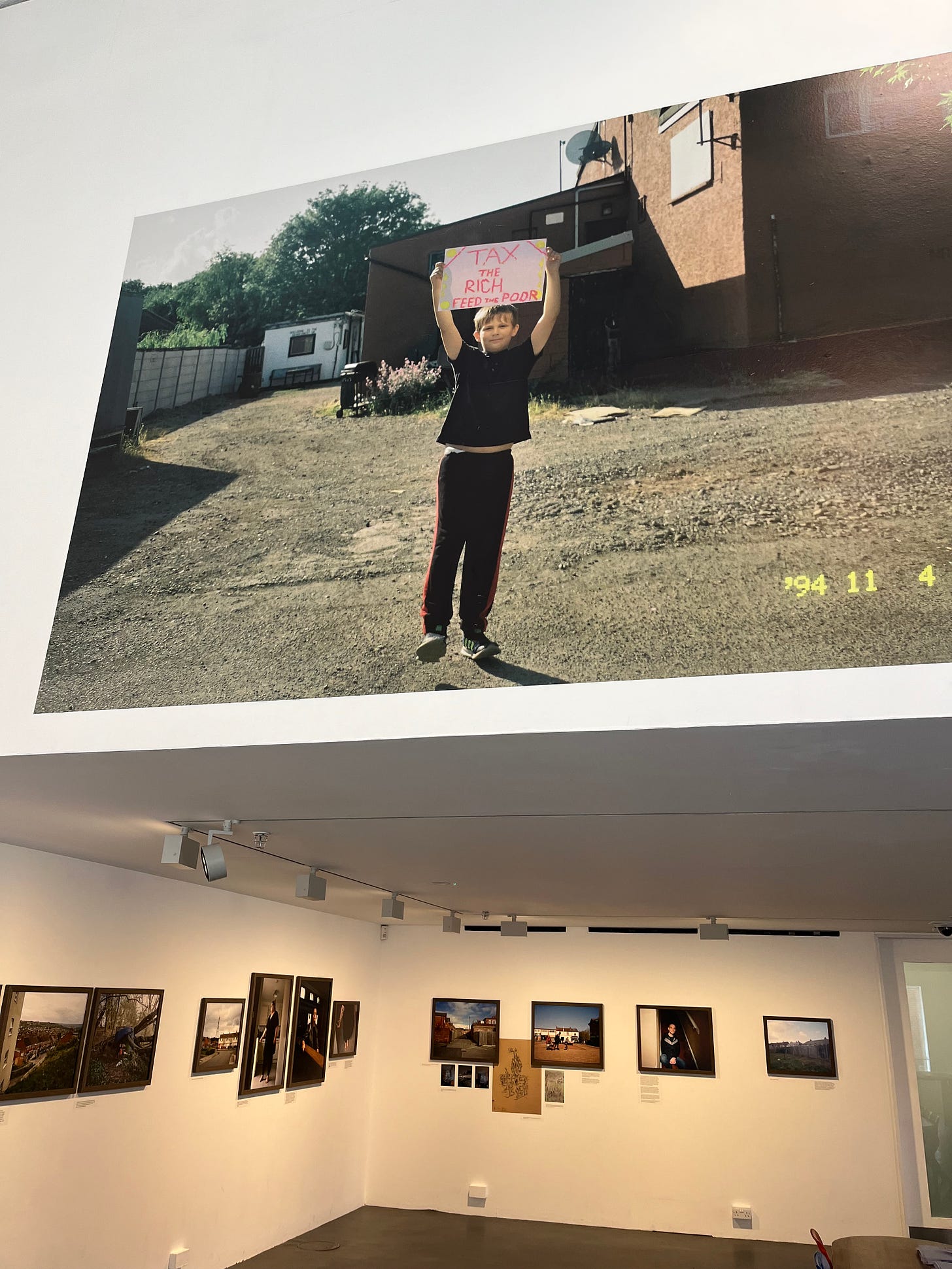 An image by Kirsty Mackay, of a boy holding a sign that says, 'Tax the rich, feed the poor.'