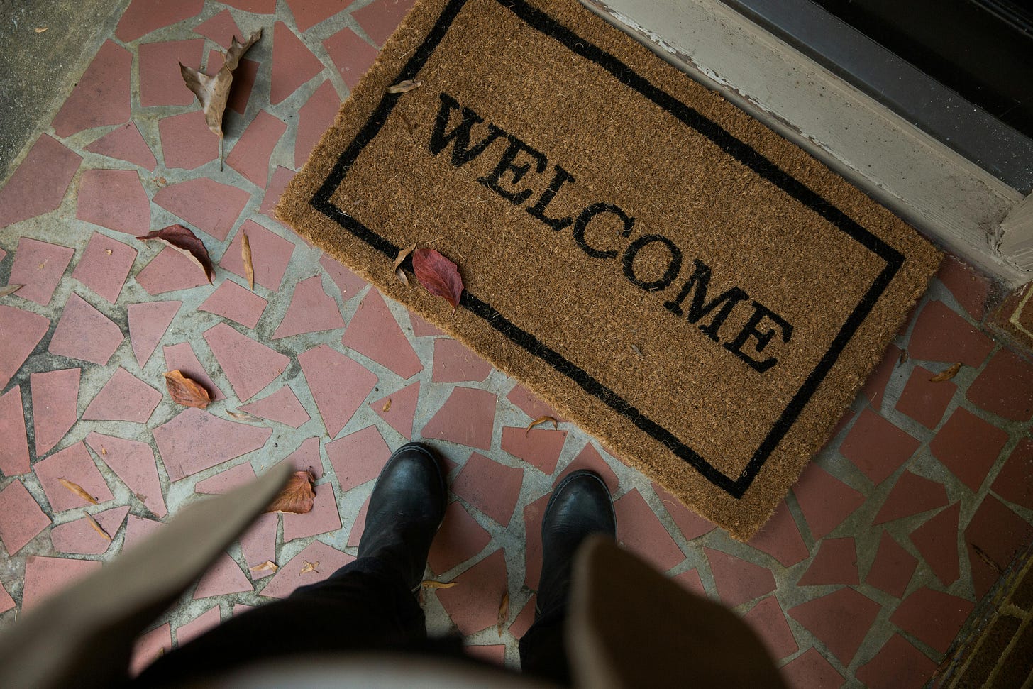 A welcome mat sits in front of a door against a tiled walkway, with two legs/feet in view.