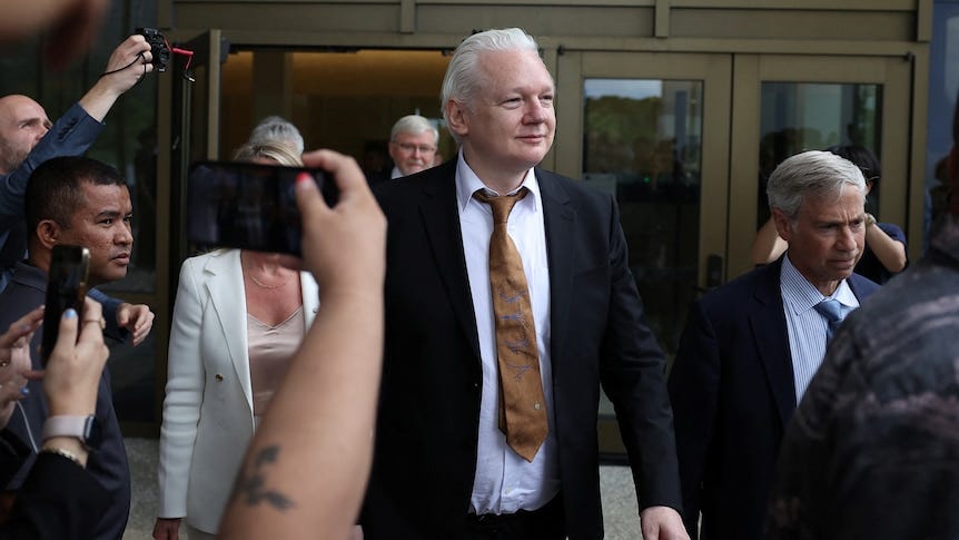 Julian Assange aux cheveux blancs portant un costume noir et une cravate marron marchant parmi une foule