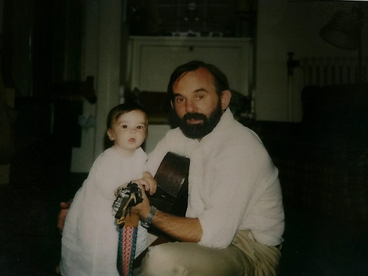 A small white child in a long pale garment stands in 3/4 view on the left. The child is holding onto the neck of a guitar being held by a man squatting on the right. He is white, too, with a beard and dark hair.