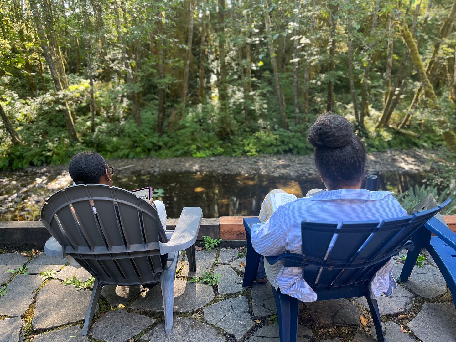 Nafissa and Jackie sit in chairs overlooking a stream in the woods as they read. Their backs are to the camera. 