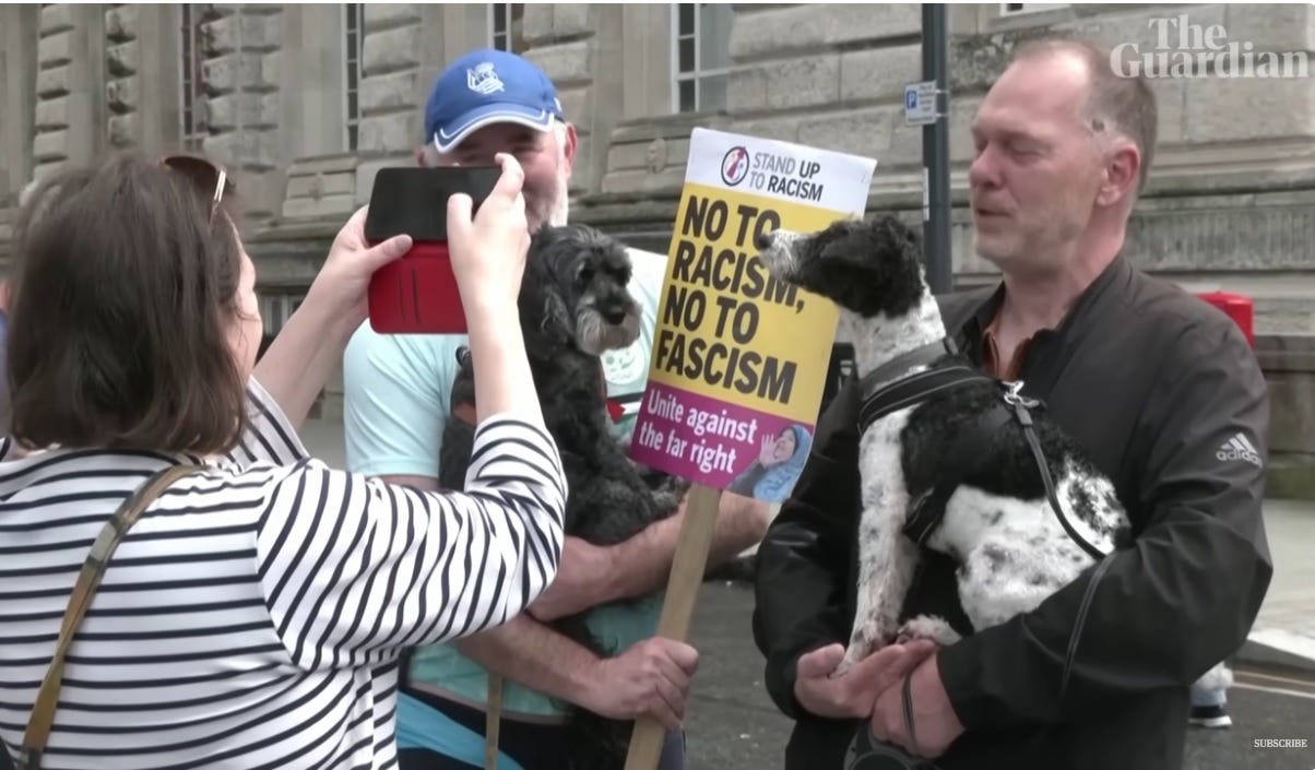 A woman uses her phone to take a photo of two fellow anti-racism marchers, each of whom holds a medium-sized dog in his arms. One man also holds a placard reading 'No to racism, no to fascism. Unite against the far right.'