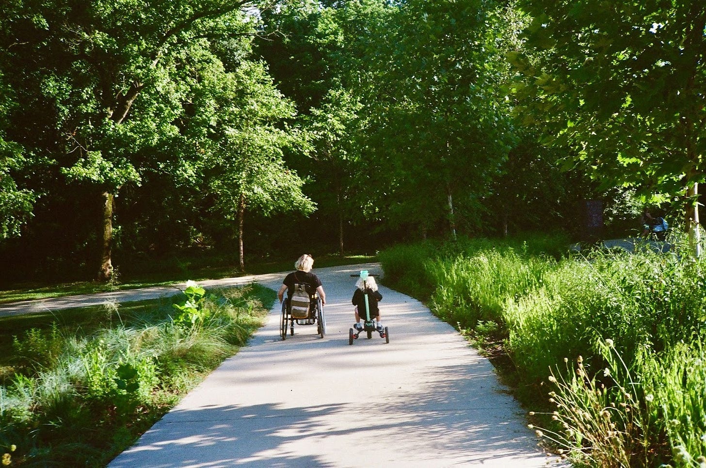 The photo captures Rebekah and her son from behind and at a little distance as they roll side-by-side down a wide, paved path surrounded by green trees and plants, she in her wheelchair, he on his tricycle. Rebekah’s head is tilted to the side, watching her son as they fly.