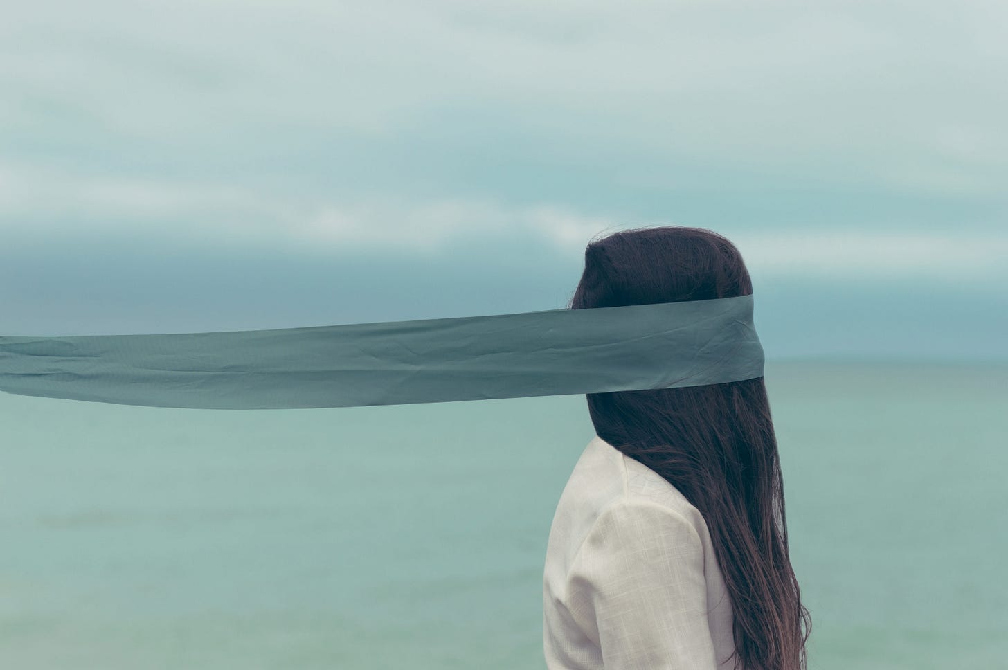 image of a long-haired person in a white shirt by the ocean on a cloudy day blindfolded by a long piece of fabric, the color of which mirrors the ocean and sky