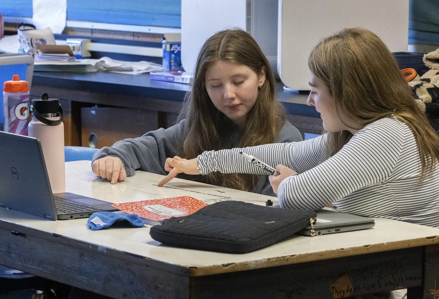 An image of two kids drawing and pointing to a number line on a desk using dry erase markers. A laptop and notebook are also visible.