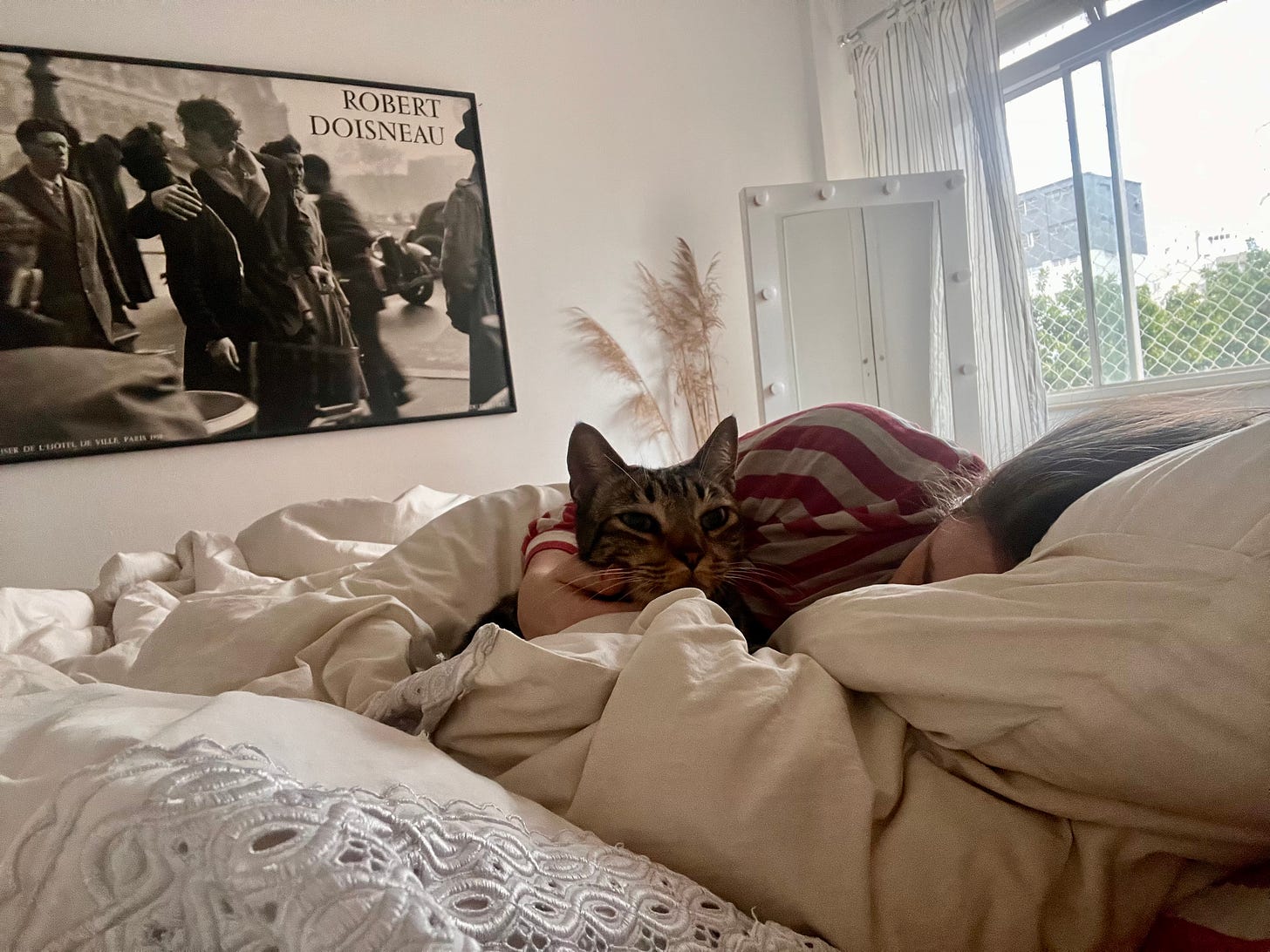 Eu deitada na cama toda branca abraçada ao gato Fortunato. Ao fundo, na parede, uma fotografia emoldurada enorme de Robert Doisnau na parede