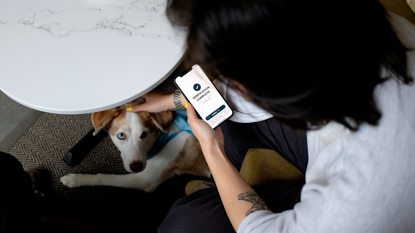 A mobile phone user viewing a Vouchsafe screen with a prominent tick, saying "verification complete". With their other hand, they pet a cute dog.