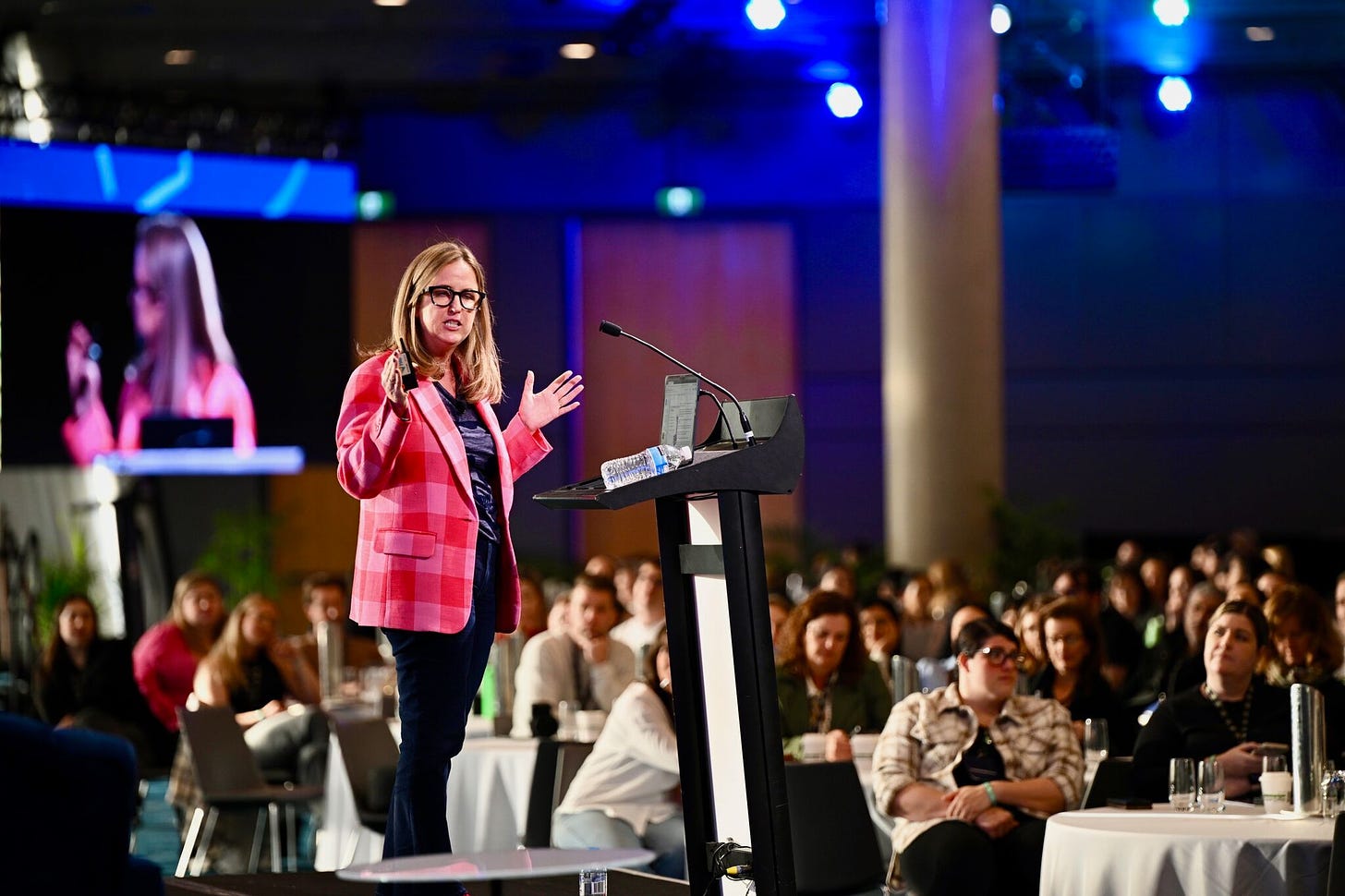 Lia Haberman standing at a podium in front of a blurry audience speaking at the State of Social conference wearing an amazing red and pink jacket from Trina Turk that manifests good fortune xo