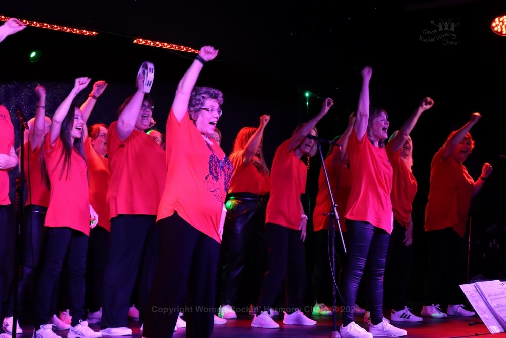 Jackie and Tanya leading Women Rockin Harmony on stage, the whole group have their right arms raised