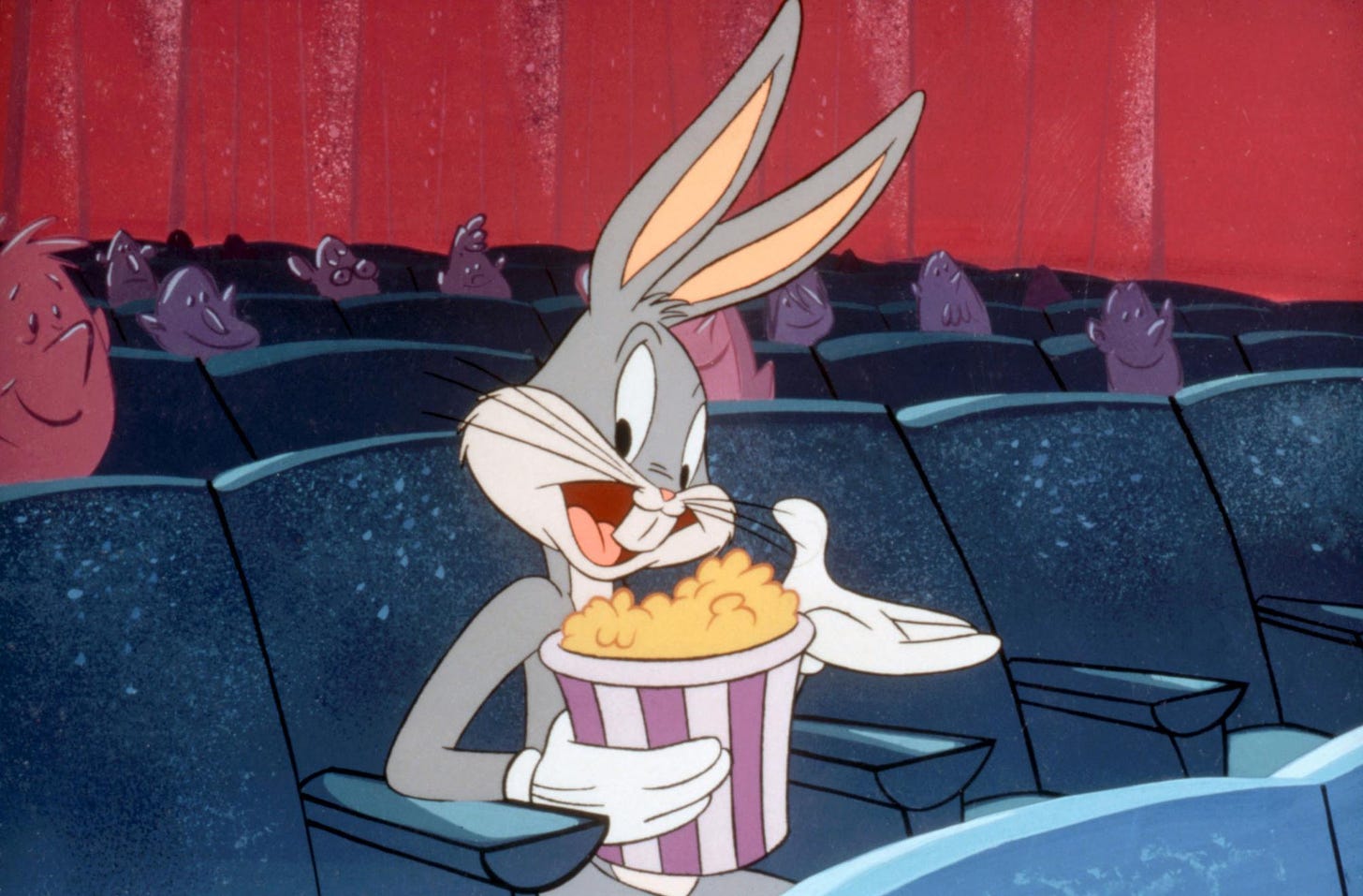 Poltrone blu del cinema con Bugs Bunny che tiene in mano un sacchetto di pop corn e ci guarda sorridendo e allargando l'altra mano. Dietro di lui spuntano qua e là delle teste rosa e viola di spettatori che guardano lo schermo. Fondale rosso.