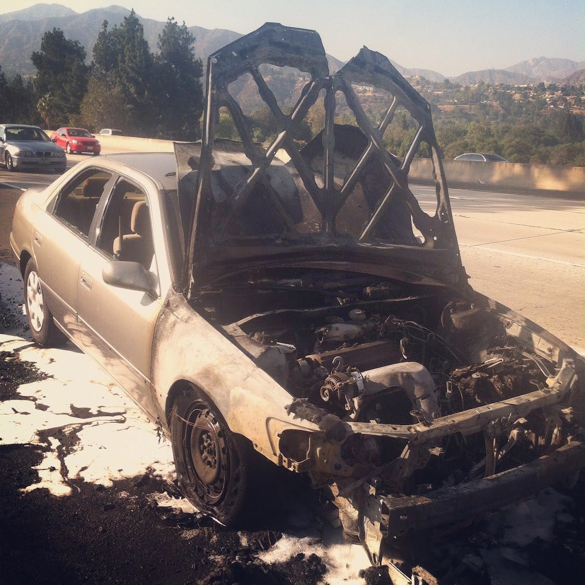 A beige 2001-ish Toyota Camry with a burnt-to-a-crisp hood and engine on the side of an LA freeway.