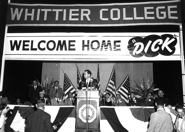 Old black and white photo of Richard Nixon in the middle distance. He stands behind a lectern on stage, head turned to his right, with five American flags behind him. Two giant banners are above him: WHITTIER COLLEGE is above WECLOME HOME "DICK"