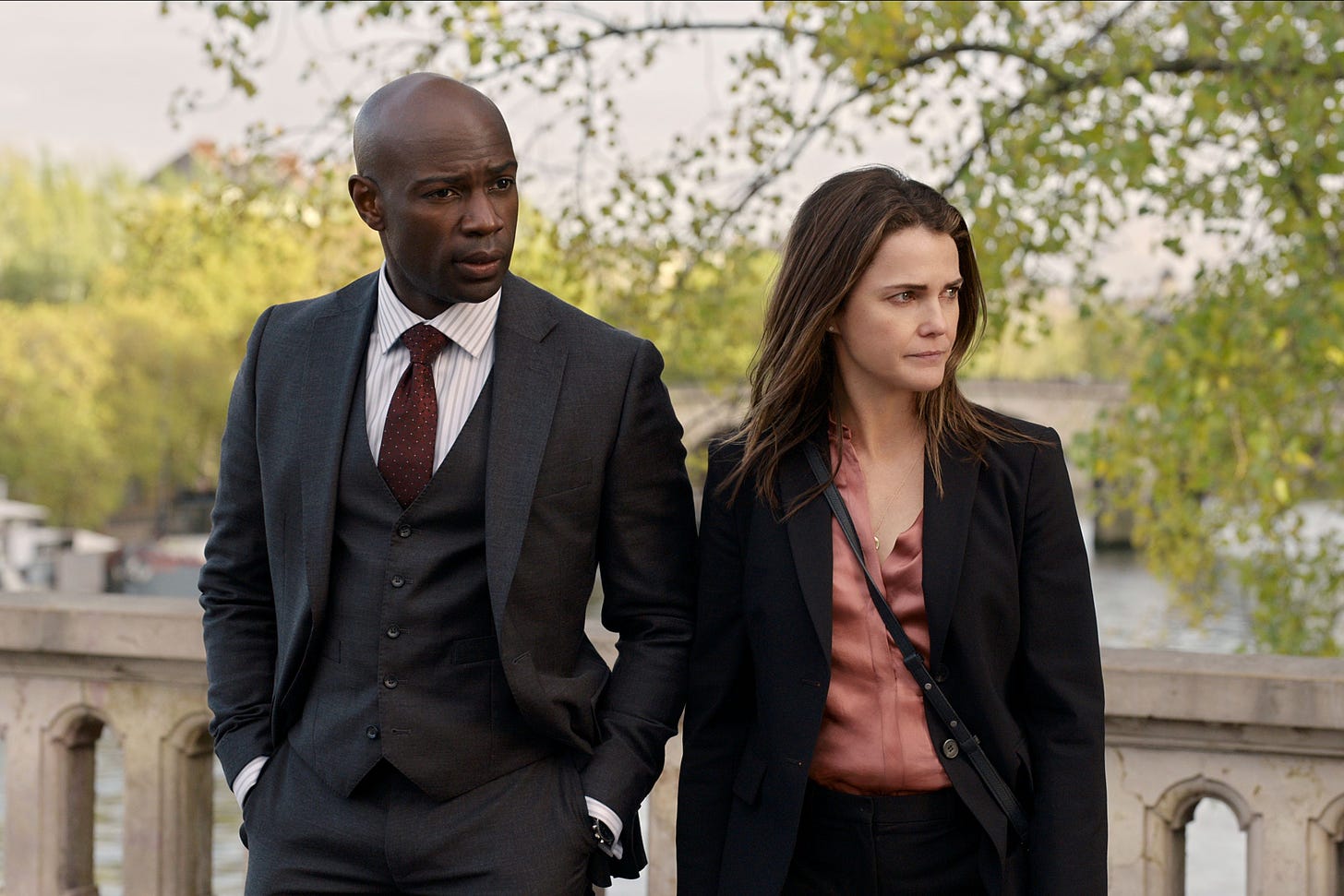 Still from the tv show The Diplomat. Two people stand on a bridge in Paris over the Seine. The man on the left is in a sharp suite and well chosen burgundy tie. Glowing black skin and shaved head. Woman on the right is in an off the rack black suit, unbrushed hair, and thinking deeply. Clearly these two are destined for some fun together.