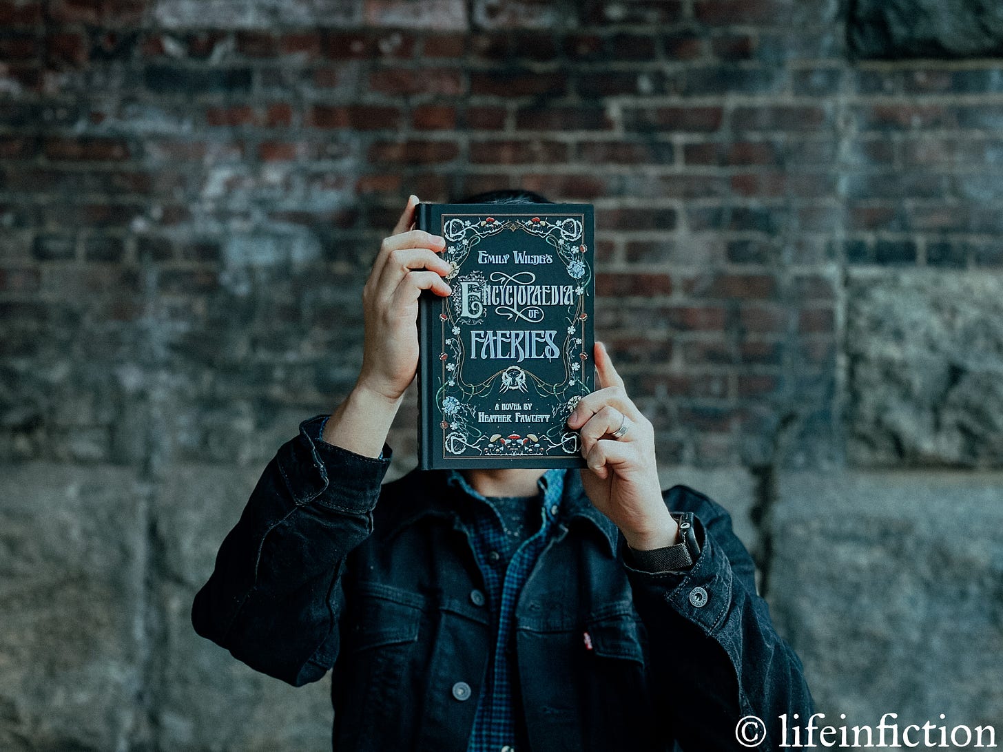 DJ holding a hardcover copy of Emily Wilde's Encyclopedia of Faeries by Heather Fawcett in front of his face. He's wearing a black denim jacket and is standing in front of a stone and brick wall.