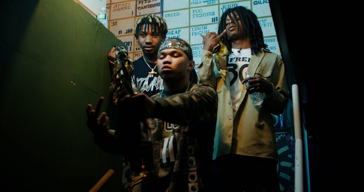 Three men on a dimly lit staircase, surrounded by posters with band names.