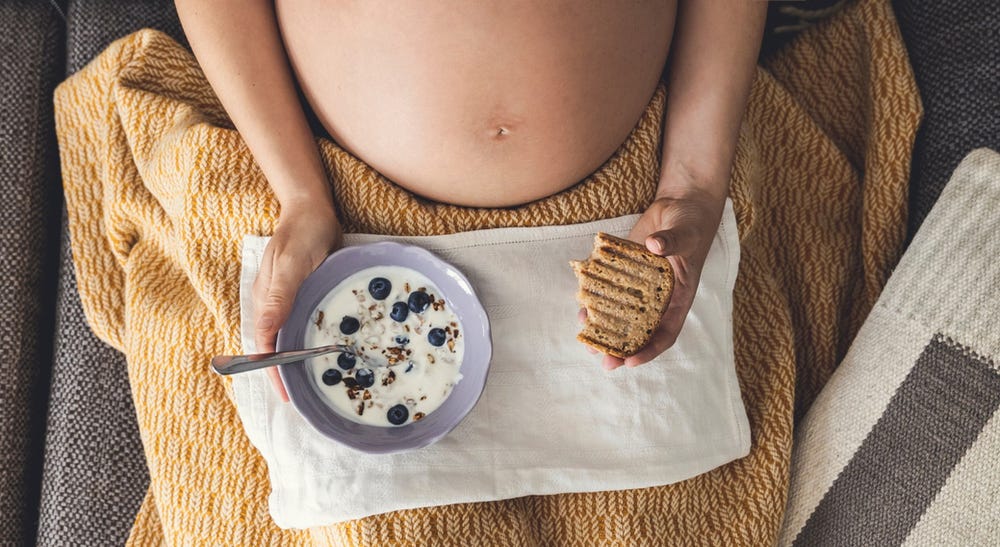 pregnant woman under a blanket with a bowl of yogurt and toast