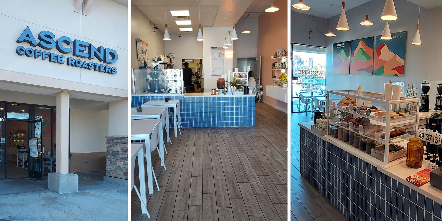 From left: The block san-serif font sign for Ascend Roasters in black against a beige wall above the entryway. A view of the cafe from the doorway. White tables on the left and a blue front tiled coffee bar at the back. The pastry case is filled to the max with fresh baked goods and coffee grinders line the counter in the background. On the wall, an abstract print of mountain silhouettes in various colors spans the wall.