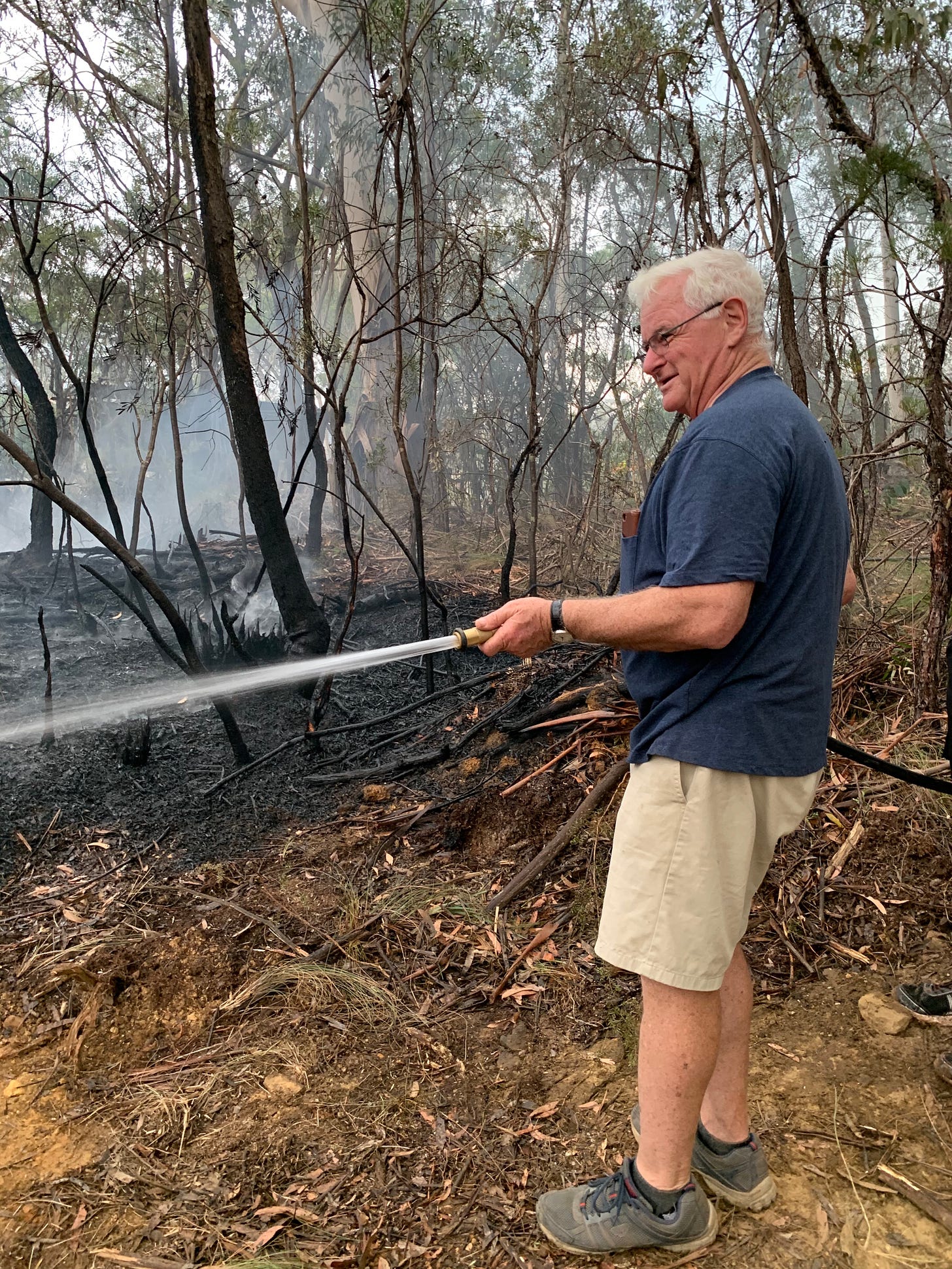 Older man in shorts and t-shirt holding a hose and watering area of burned smoking ground.