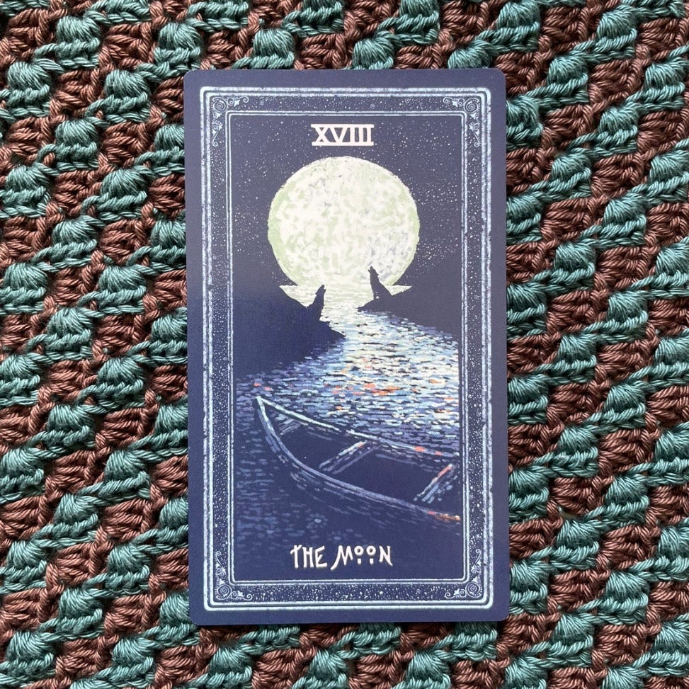A tarot card sits on a turquoise and brown striped crocheted cloth. It is a dark blue with a sliver edging and a picture of a full moon reflecting down a meandering river. Two wolves are silhouetted against the moon, sitting on opposite river banks and howling. In the foreground is an empty wooden boat. At the top of the card in capitals are the roman numerals XVIII and at the bottom it says THE MOON.