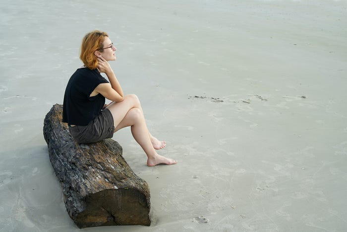 white woman wearing glasses with short hair sitting on a log on a sandy beach