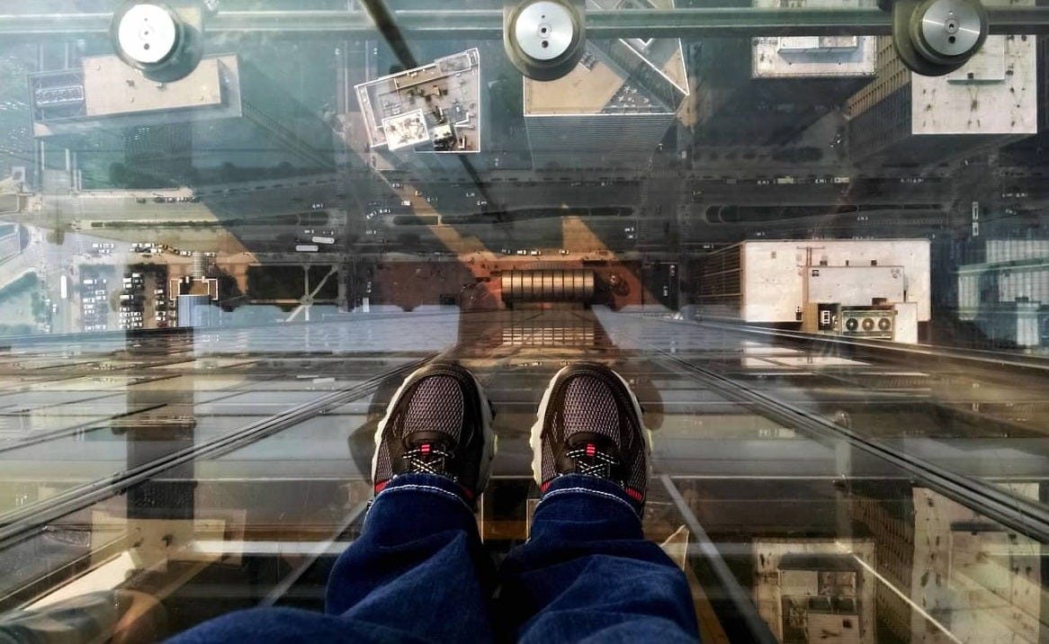 A few of two sneakered feet standing on a glass platform high above Chicago, looking straight down at the rooftops and streets below