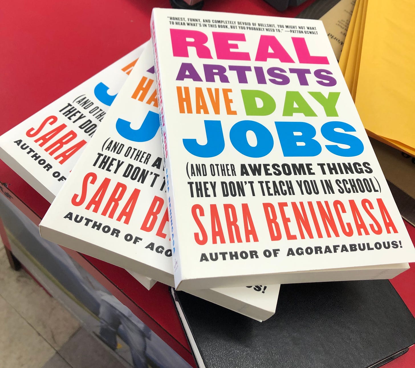 Three copies of "Real Artists Have Day Jobs" by Sara Benincasa, atop a red bookshop counter