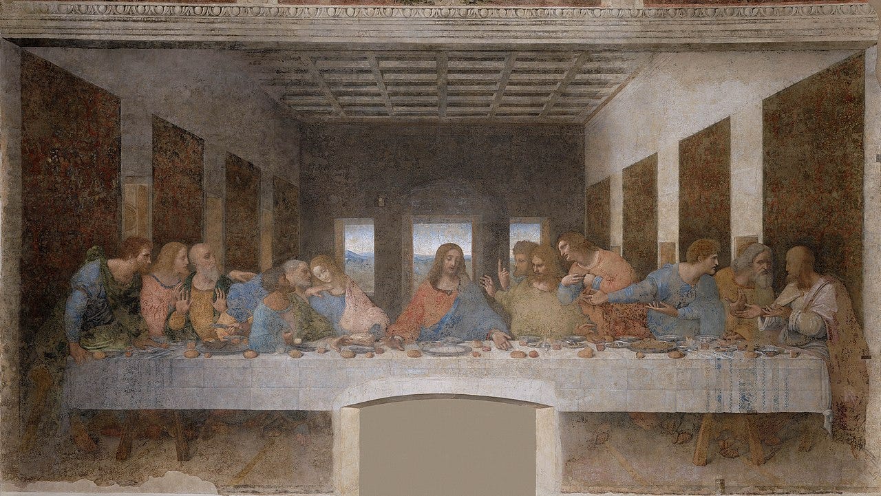 Leonardo da Vinci’s The Last Supper, circa 1495–1498. A painting, roughly twice as wide as it is tall. In the lower half, most of the composition is taken up by a long, horizontal table with Jesus seated at the center with his twelve apostles to his left and right. The figures are all facing the viewer. They are indoors, and most of the upper half of the images is devoted to precise, geometric architectural details such as walls, the ceiling, and, in the background, three vertical windows with Jesus framed in the center window. A green landscape in the far distance is visible through the windows. The table is set with a white tablecloth, wine glasses, water pitchers, bowls, bread, fish, and fruits. The group is depicted at the moment Jesus tells them one of them will betray him. Each apostle reacts with different gestures and expressions.