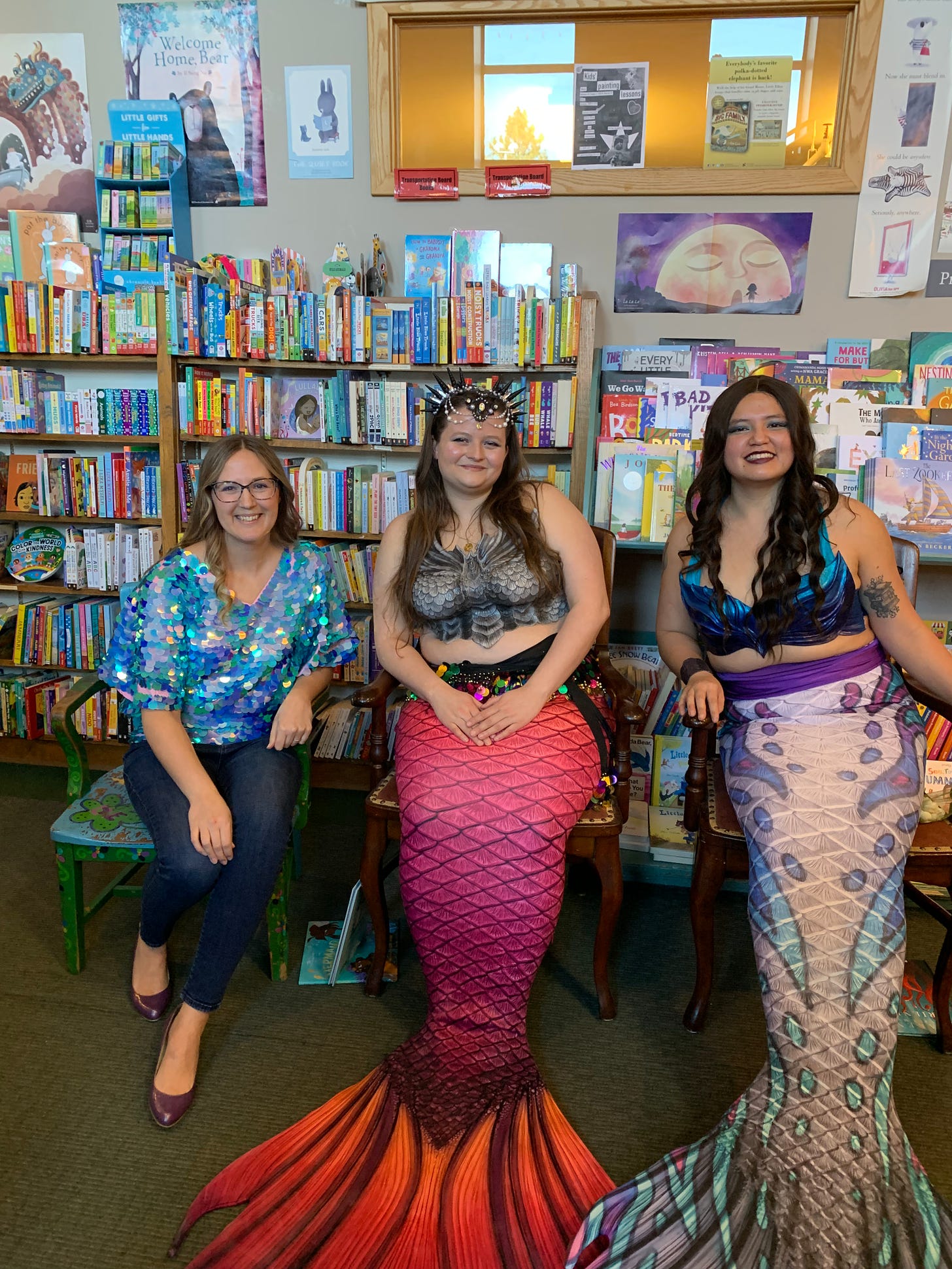 Eija wearing a blue sequin scales shirt sitting next to two mermaids in the children’s section of Auntie’s Bookstore